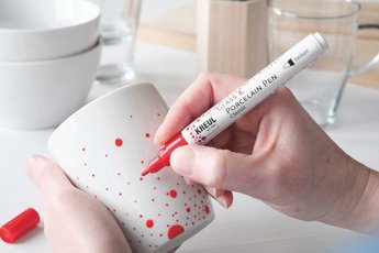 Hand holding a red KREUL Glass & Porcelain Pen demonstrating how to apply decorative designs with red dots on a white porcela
