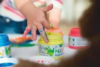 MUCKI Finger Paint white 150 ml jar with colorful children’s artwork in the background.